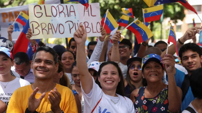 Un grupo de personas aglomeradas sonríen mientras ondean pequeñas banderas de Venezuela.