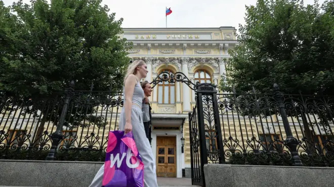Dwie kobiety przechodzą obok bladożółtego budynku z rosyjskim napisem „Bank Rosji” i rosyjską flagą w kolorach białym, niebieskim i czerwonym.