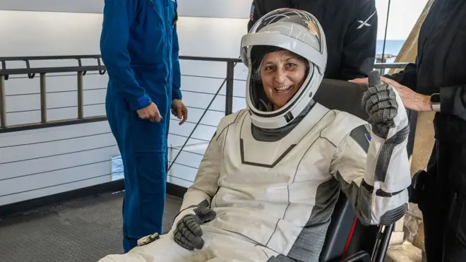 Suni Williams smile as she comot di capsule for inside one white space suit and helmet, two assistants wey wear black dress help her