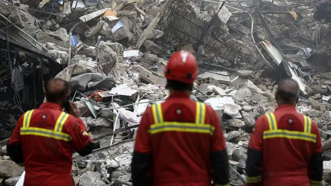 Three men in red emergency service worker uniforms survey damage from a strike on Tehran, Iran. Photo: 23 March 2026. 