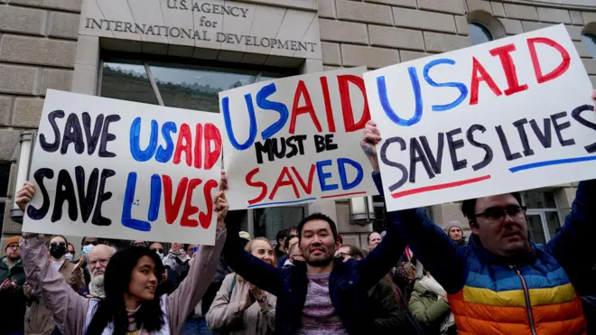 Люди митингуют у здания USAID, на переднем плане три человека с плакатами. На них написано «Спасите USAID, спасите жизни», «USAID должен быть спасен» и «USAID спасает жизни».