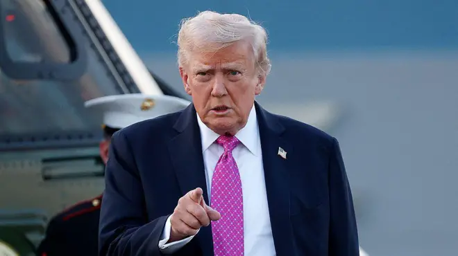 د امریکا ولسمشر ډونلډ ټرمپ په درېشۍ کې ولاړ دی چې ګلابي رنګه خالداره نېټکټايي یې اچولې او ګوته یې مخامخ په اشاره نیولې ده.