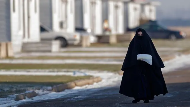 Tamamen siyah giyinmiş, saçları örtülü ve sadece gözleri görünen bir kadın, oluklarda kar görünen boş bir caddede yürüyor.