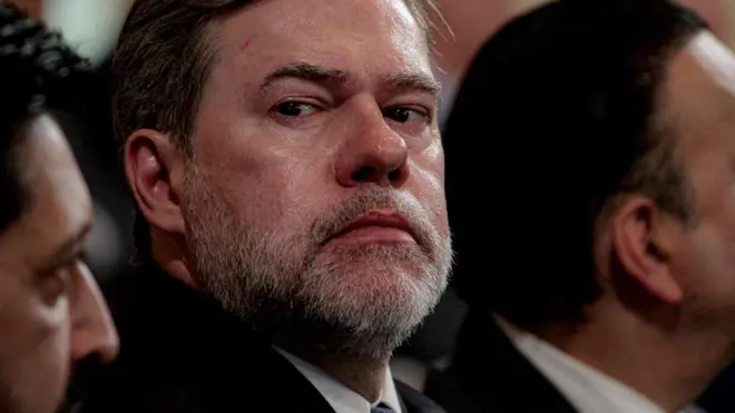 O ministro José Dias Toffoli, do STF, um homem de meia-idade, com barba grisalha e expressão séria, olha de lado. Ele veste terno escuro e camisa social, e está sentado entre outras pessoas, que aparecem desfocadas ao redor. A iluminação destaca seu rosto.