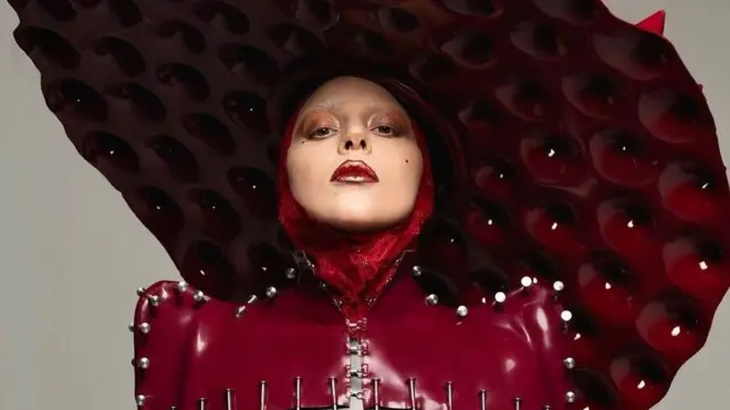Lady Gaga usa uma roupa de vinyl vermelho-sangue, costurada com pregos e coberta por um chapéu enorme em forma de disco