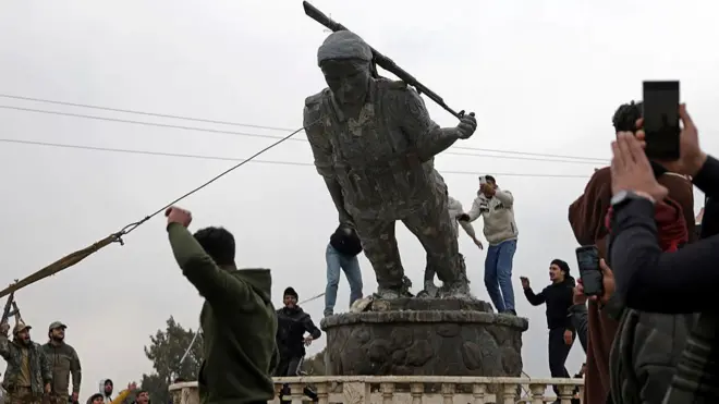 Çok sayıda kişi bir heykeli yıkmak için birlikte çalışıyor. Heykelin bir kürt kadın savaşçıya ait olduğunu biliyoruz. Heykel, uzun namlulu silah tutan bir kadını temsil ediyor.