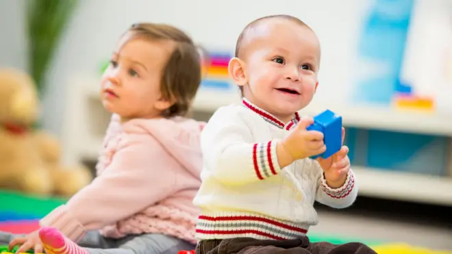 Dois bebês sentados no chãoapostas desportivas bonus gratisuma salaapostas desportivas bonus gratisjogos, um olhando para a frente e segurando um brinquedo, e o outro ao fundo, olhando para longe 