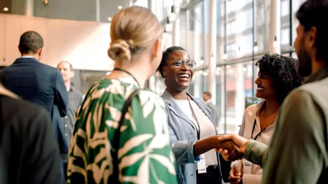 Mulheres e homens com roupas profissionais e crachás de empresa sorrindo e se cumprimentando, em uma roda de conversa