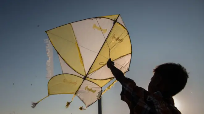 A silhouette of a young boy holding a kite up against the sky, with industrial buildings in the background