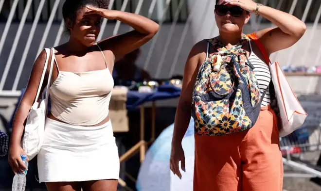 Duas mulheres cobrem os rostos com as mãos, tentando se proteger do sol, ambas vestem blusas de alcinhas e carregam bolsas e mochila