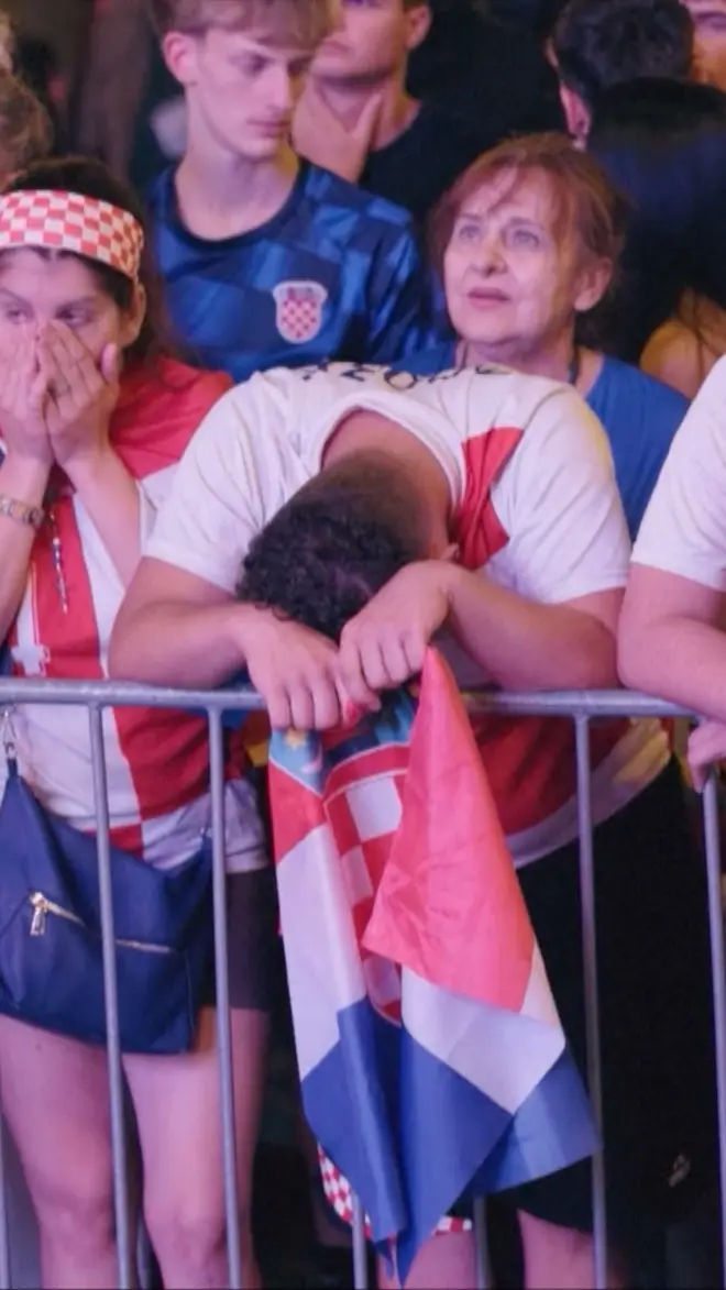 Navijač Hrvatske stoji naslonjen na ogradu, spuštene glave i držeći zastavu Hrvatske u rukama.