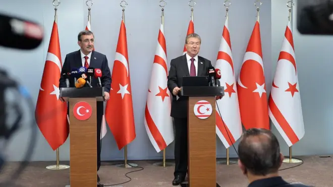 Yılmaz ve Üstel kürsüden konuşuyor. Arkalarında Türk ve Kuzey Kıbrıs bayrakları var.