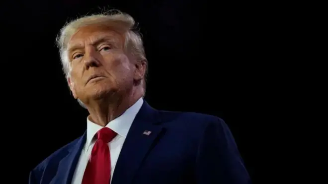 Perezida Donald TRump yambaye ikoti yirabura, ishati igomba kwera n'ikaruvati itukura, ariko araba muri camera