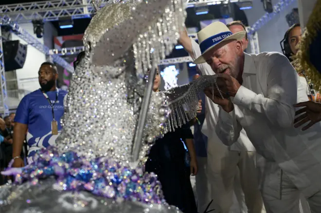 De branco e chapéu panamá, Lula cumprimenta porta-bandeira da Acadêmicos de Niterói durante desfile, beijando sua mão. Ela usa roupa azul e branca com muito brilho.
