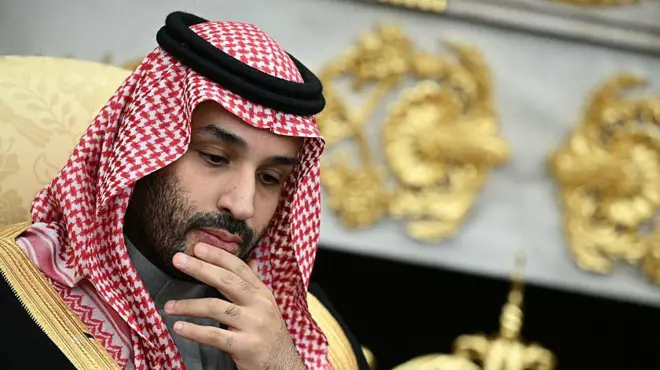 He is rubbing his chin, thinking. He is wearing traditional Arab clothing, with the red and white head dress. He is wearing a black robe with gold edges.