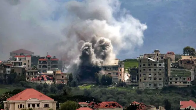 Kırsal bir yerleşim yeri gibi görünen bir yerde yüksek binaların arasında dumanlar yükseliyor. 