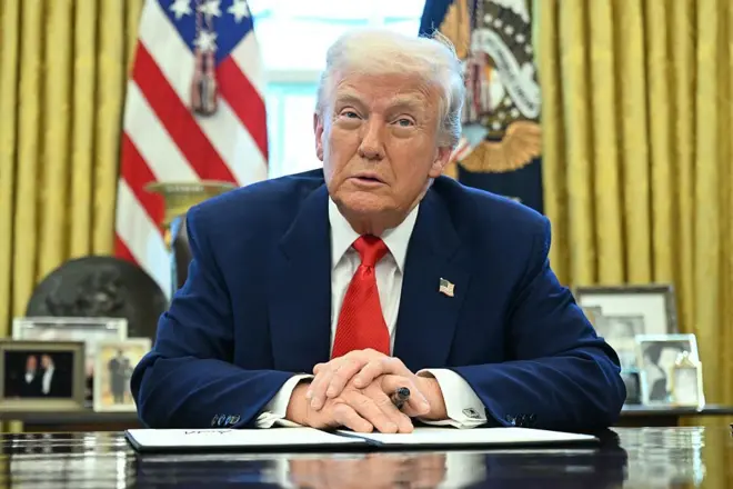 Trump arkasında Amerikan bayrağı ve başkanlık arması ile masada oturmuş, elinde bir kalem ile önündeki kağıt üzerinde ellerini kavuşturmuş