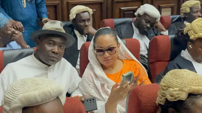 Natasha and husband holding phone while some lawyers dey background
