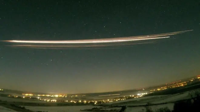 A photograph using 30-second exposure showing the Falcon 9 upper stage re-entering the atmosphere above Berlin, Germany. A long streak of bright yellow and white is seen in the dark night sky, above a landscape of dark field and bright lights from the city.