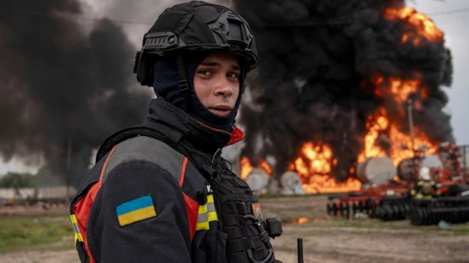 Member of di emergency services for north-eastern Sumy, wey wear high vis wit di Ukraine flag dem sew for im arm and combat helmet and wetin dem call balaclava, as im look at di camera standing in front of gas depot wey don dey burn near di Russian border