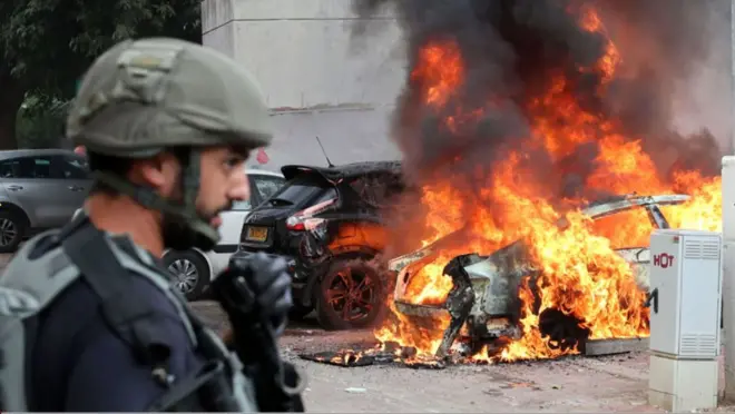 একটি গাড়িতে আগুন জ্বলছে, তাকিয়ে আছে একজন ইসরায়েলি সৈনিক