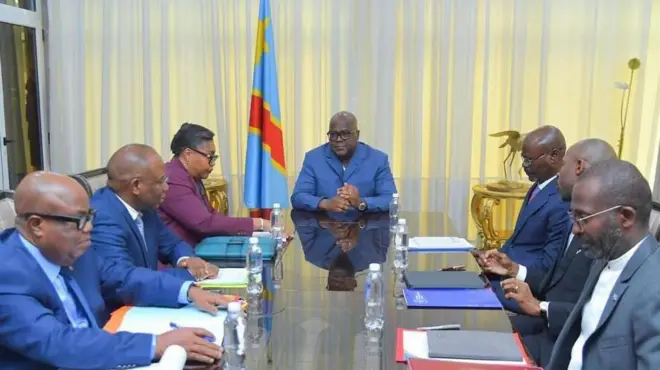 Perezida wa DR Congo Félix Tshisekedi yicaye mu nama akikijwe ku meza n'abategetsi bakuru barimo na Minisitiri w'intebe Judith Suminwa. Inyuma ya Tshisekedi hari igitambaro cyo gukingira idirishya cy'ibara rya khaki rijya kuba umuhondo hamwe n'ibendera rya DR Congo ry'ubururu, umuhondo n'umutuku. Ifoto yatangajwe n'ibiro bya Perezida Tshisekedi, ku wa kane, ku itariki ya 11 Ukuboza (11) mu mwaka wa 2025.