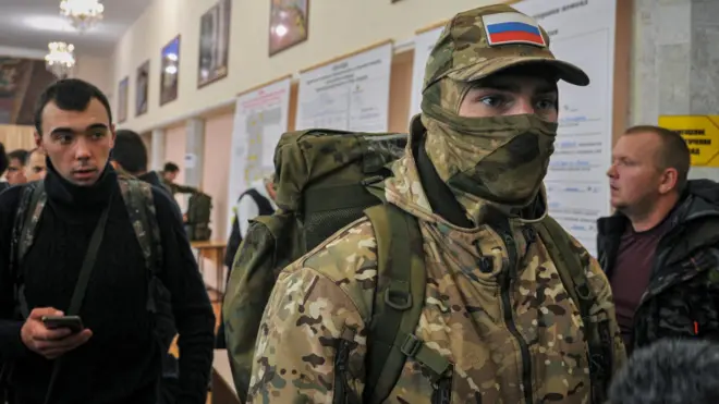Мужчина в камуфляже, маске и с рюкзаком с российскими опознавательными знаками в окружении других мужчин в штатском (26 сентября 2022 года)