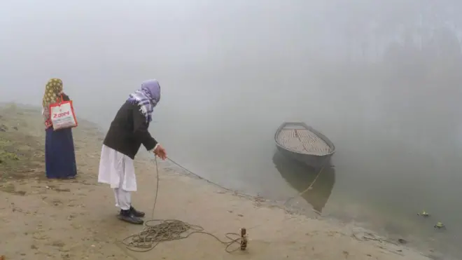 একজন নারী ও একজন বয়স্ক পুরুষ নদীর পাড়ে দাঁড়িয়ে আছে। বয়স্ক লোকটি ঘাটে বাঁধা নৌকাকে তীরে ভেড়ানোর চেষ্টা করছেন।