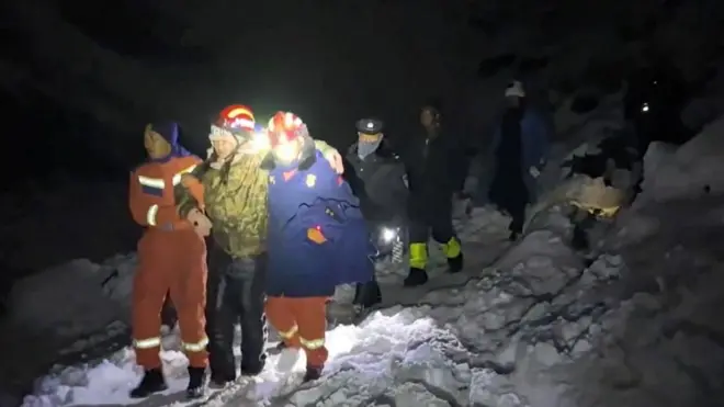 Uma pessoa sendo levada por dois homens que usam roupas de resgate e caminham na neve 