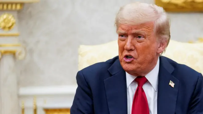 Trump is speaking, seated in the Oval Office. He wears a navy suit and red tie.