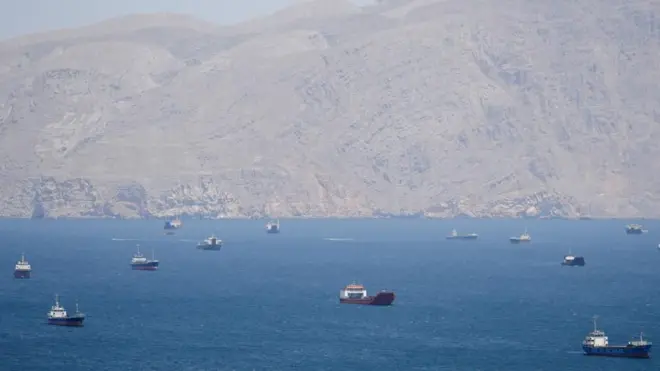 Cargo ships and small vessels scattered across deep blue sea, with hazy rocky mountains rising along distant coastline under sky