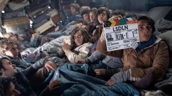 El protagonista de la película, Enzo Vogrincic, sostiene una claqueta listo para rodar una escena y detrás están sus compañeros de elenco dentro del fuselaje del avión.