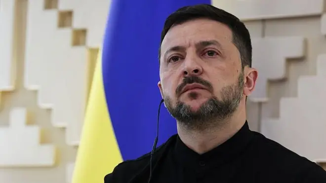 Perezida wa Ukraine Volodymyr Zelensky ari iruhande y'ibendera ry'umuhondo n'ubururu rya Ukraine. Yambaye umupira umeze nk'ishati w'umukara, afite n'igikoresho cyo kumvisha mu gutwi kumwe. 