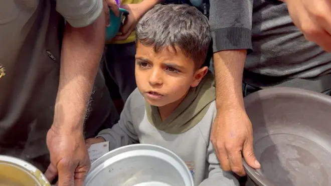 A young boy, holding a metal bowl, standing in a crowd waiting for food