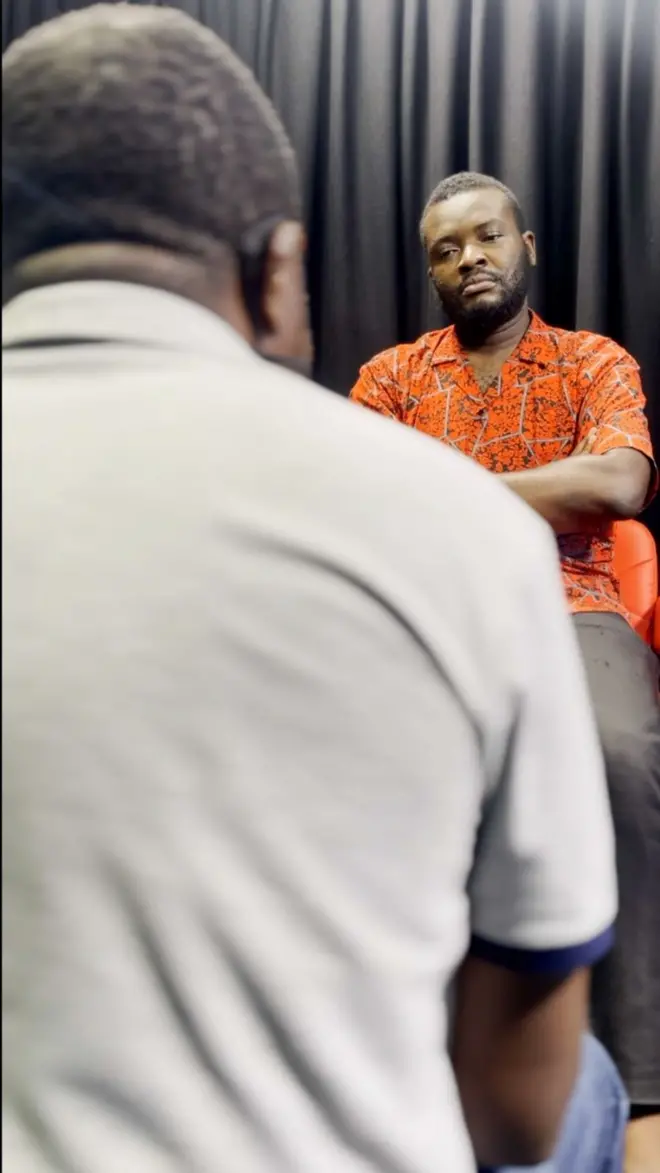 Two men sitting down - one is facing the camera, while the other is backing it