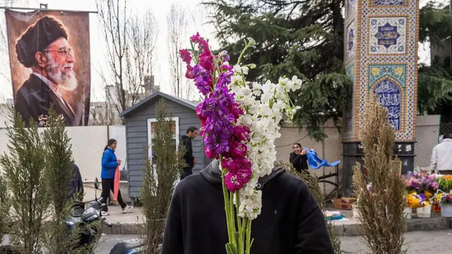A flower vendor holds purple and white flowers over his face as people shop in preparation for Nowruz celebrations on 19 March 2026 in Tehran, Iran. Behind him on the left, a poster of former Supreme Leader Ayatollah Ali Khamenei, who was killed in the first wave of US-Israeli strikes, is displayed. A few people walk behind the vendor. 