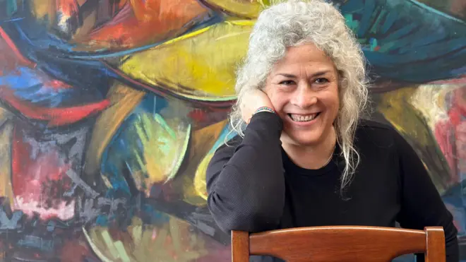 Olga Montero Rose sonríe sentada en una silla de madera con el respaldo hacia el frente y su codo derecho apoyado en él y su mano derecha sobre su rostro. Detrás hay una pintura.