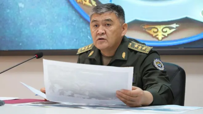 Седой Камчыбек Ташиев сидит за столом и рассматривает фотографии. Он одет в зеленую военную форму