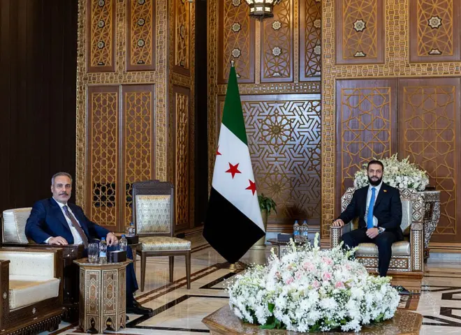 hakan fidan ve ahmed şara, suriye'deki bir salonda kameralara poz verirken