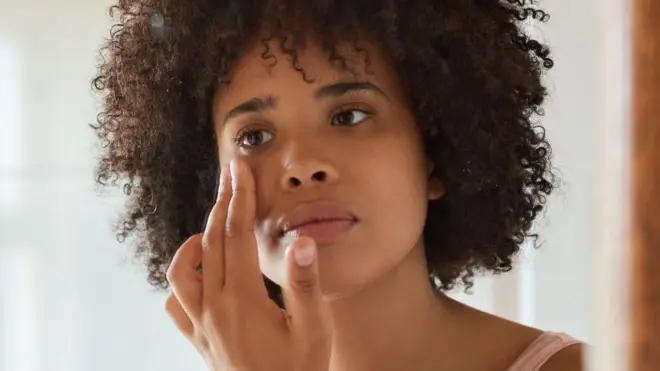 Una joven de cabello oscuro se mira al espejo, tocándose la piel cerca del ojo.