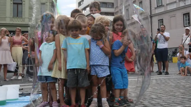 Imagem mostra crianças dentrouma bolhasabão observadas por grupoadultos,uma praça na Europa