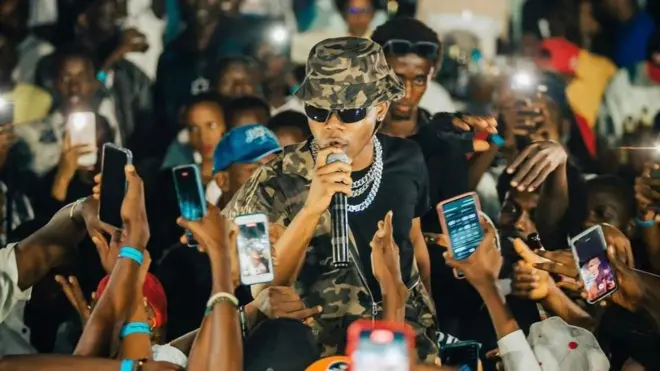 Umuhanzi w’umugabo yambaye amarori n’inkofero arimo kuvugira muri ‘micro’. Akikujwe n’abantu benshi, bamwe bafite telefone ngendanwa bariko baramufotora.