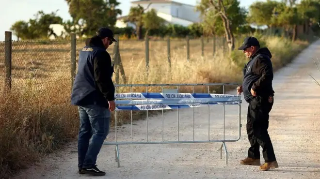 Двое полицейских несут перемотанный полицейской лентой кусок ограждения, чтобы перегородить грунтовую дорогу. Вдалеке виден частный дом.