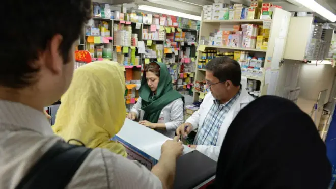 پشت پیشخوان یک داروخانه که جند نفر برای گرفتن دارو صف کشیده‌اند.