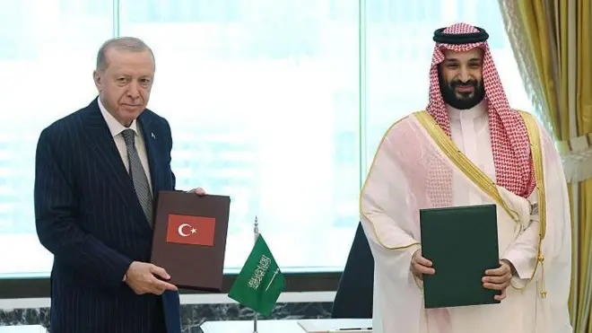 Takım elbiseli Cumhurbaşkanı Erdoğan elinde türk bayraklı bir dosya tutuyor. Yanındaki beyaz yerel elbiseli sakallı prens de yeşil renkli bir dosya tutuyor ve gülüyor.