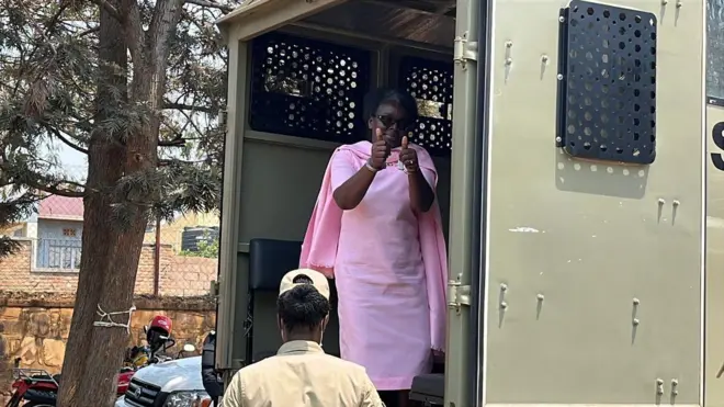 Victoire Ingabire ahagaze mu muryango ufunguye w'inyuma w'imodoka yo mu bwoko bw'ivani itwara imfungwa, arimo kureba muri 'camera'. Akoze ikimenyetso n'ibikumwe bibiri, ari ku mapingu. Yambaye ikanzu n'umwitero ku ijosi no ku rutugu, byose biri mu ibara ry'iroza riranga imfungwa. Imbere ye hahagaze umucungagereza wambaye imyenda ya khaki n'ingofero, ateye ibitugu 'camera'. Ifoto yo ku itariki ya 26 Kanama (8) mu 2025. 