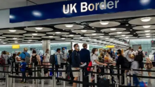 Aworan yii ṣafihan awọn alawọ funfun lẹnu ibode abawọle UK. Pupọ wọn pọn baagi sẹyin, wọn si fi ibomu bo imu wọn. Ina tan lati oke abawọle naa, bẹẹ ni wọn fi awọn okun dudu dá aaye ti awọn arinrinajo to fẹẹ wọle naa duro si. Akọle to kede ẹnu ibode naa han loke pẹlu.