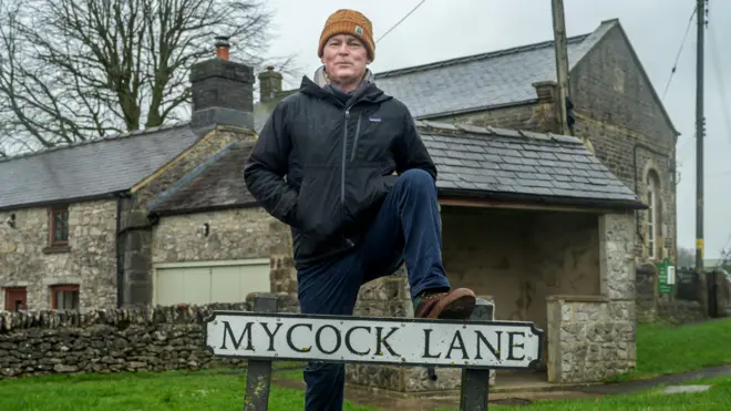 A smiling man in a raincoat and yellow hat stand with one foot resting on a road sign that says "Mycock Lane"