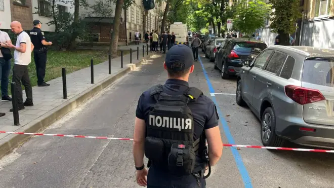 На передньому плані поліціянт у бронежилеті за огороджувальною стрічкою. Далі – група правоохоронців і поліцейська автівка