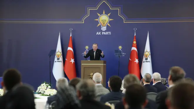 Erdoğan AKP logolu kürsüden katılımcılara sesleniyor kürsünün sağ ve sol yanında Türkiye ve AKP bayrakları var.